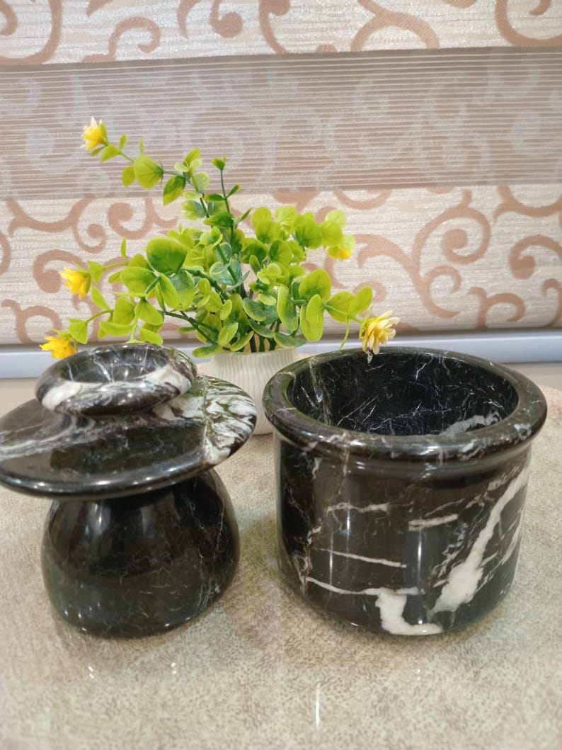 May include: Two black marble containers with white veining. One is a small, lidded jar, and the other is a cylindrical pot. The containers are on a light-colored surface, with a small plant in the background.