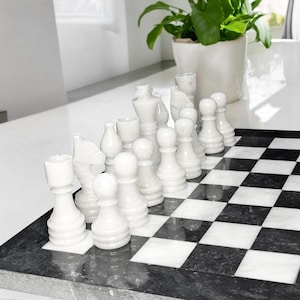 Pu&ograve; includere: Un set di scacchi con pezzi in pietra bianca lucida disposti su una scacchiera quadrata con un motivo a scacchi bianco e nero a contrasto. Il set include varie figure intagliate come pedoni, cavalli e un re, realizzate in pietra liscia.