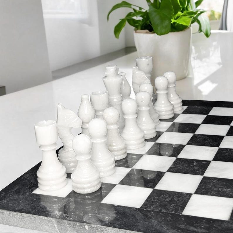Pu&ograve; includere: Un set di scacchi con pezzi in pietra bianca lucida disposti su una scacchiera quadrata con un motivo a scacchi bianco e nero a contrasto. Il set include varie figure intagliate come pedoni, cavalli e un re, realizzate in pietra liscia.