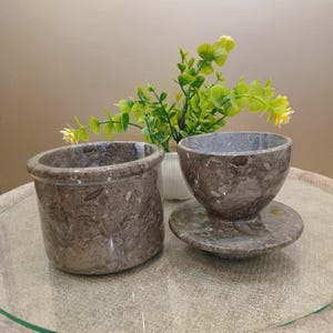 May include: Two brown marble containers. One is a small, round pot with a thick rim. The other is a small bowl with a matching saucer. The marble has a speckled pattern. A small plant with green leaves and yellow flowers is in the background.