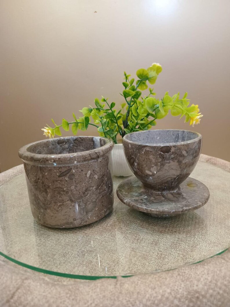 May include: Two brown marble containers. One is a small, round pot with a thick rim. The other is a small bowl with a matching saucer. The marble has a speckled pattern. A small plant with green leaves and yellow flowers is in the background.