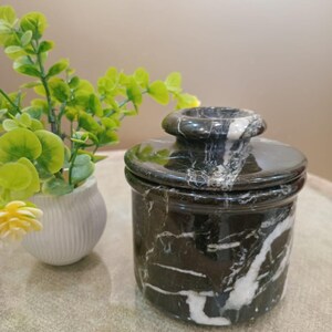 May include: A black marble butter dish with white veining. The dish has a round base and a matching lid with a small handle. A small white vase with green and yellow plants is in the background.