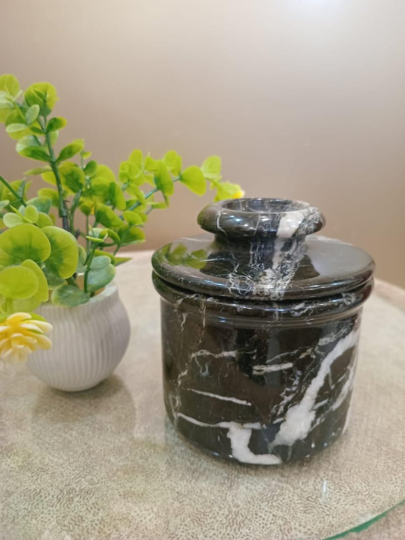 May include: A black marble butter dish with white veining. The dish has a round base and a matching lid with a small handle. A small white vase with green and yellow plants is in the background.