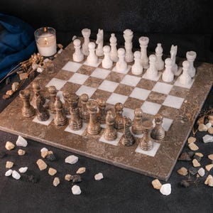 May include: A marble chess set with white and brown pieces on a checkered board. The board is set on a brown marble base. The chess pieces are arranged for a game. A lit candle and decorative stones are in the background.