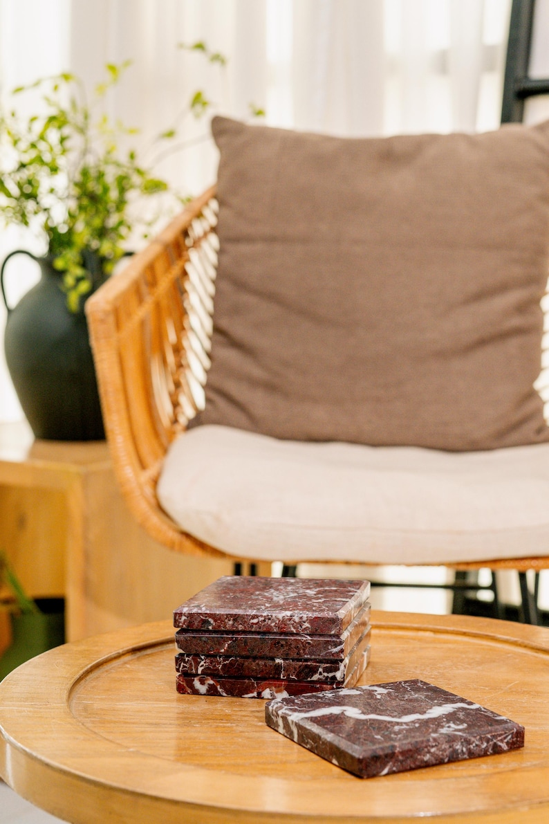 Pu&ograve; includere: Un set di cinque sottobicchieri quadrati in marmo rosso scuro con venature bianche prominenti. Quattro sottobicchieri sono impilati in una colonna verticale, mentre un singolo sottobicchiere &egrave; appoggiato in piano sulla superficie davanti alla pila, mostrando la texture della pietra.