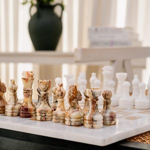 Pode incluir: Um jogo de xadrez com um tabuleiro quadrado de pedra branca com casas claras e escuras alternadas. Um conjunto de peças é feito de pedra multicolorida com faixas castanhas e creme, enquanto o outro conjunto é de pedra branca sólida.