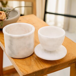 May include: Two white marble vessels sit on a wooden surface. One is a cylindrical container, and the other is a small cup on a saucer. A bowl with flowers is in the background. The items are likely for home decor.