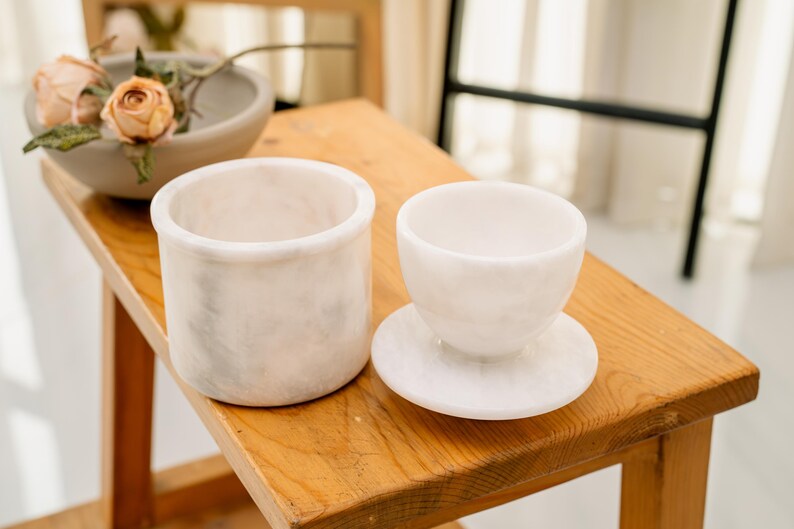 May include: Two white marble vessels sit on a wooden surface. One is a cylindrical container, and the other is a small cup on a saucer. A bowl with flowers is in the background. The items are likely for home decor.