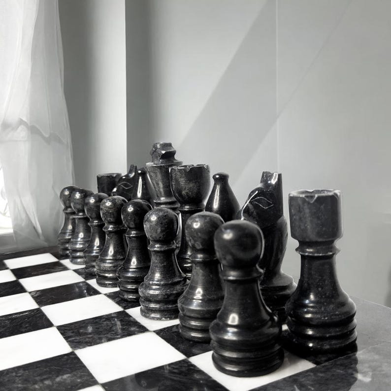 Pu&ograve; includere: Un set di pezzi degli scacchi in marmo nero disposti su una scacchiera abbinata in marmo bianco e nero. I pezzi includono pedoni, torri, cavalli, alfieri e un re, tutti caratterizzati da una consistenza in pietra liscia e lucida con venature.