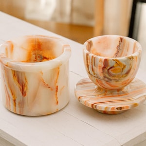 Könnte beinhalten: Zwei dekorative Onyxstücke. Eines ist ein zylindrischer Aschenbecher mit abgerundetem Rand. Das andere ist eine kleine Tasse mit Untertasse. Beide Teile haben eine cremefarbene Basis mit orangefarbenen und braunen Adern.
