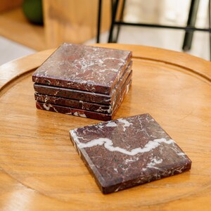 Pu&ograve; includere: Un set di cinque sottobicchieri quadrati in marmo rosso scuro lucido con venature bianche prominenti. Quattro sottobicchieri sono impilati in una colonna verticale, mentre un singolo sottobicchiere &egrave; appoggiato in piano davanti alla pila.