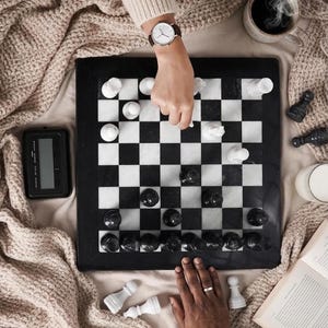 Pu&ograve; includere: Una scacchiera quadrata in marmo con caselle alternate bianche e nere, completa di un set di pezzi degli scacchi in pietra abbinati. Un orologio digitale per scacchi si trova sulla sinistra, accanto a un libro e una tazza di caff&egrave;.