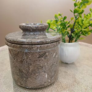May include: A brown marble butter dish with a lid. The dish has a cylindrical shape with a rounded top and a small knob on the lid. The marble has a speckled pattern. A small white vase with green plants is in the background.