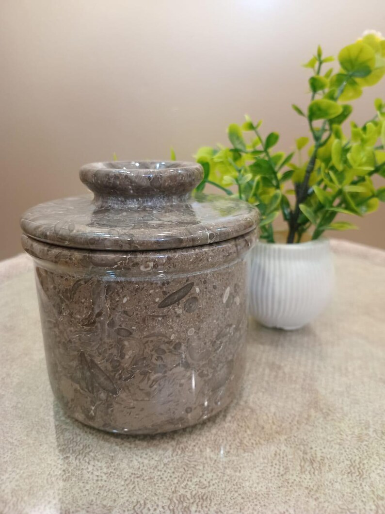 May include: A brown marble butter dish with a lid. The dish has a cylindrical shape with a rounded top and a small knob on the lid. The marble has a speckled pattern. A small white vase with green plants is in the background.