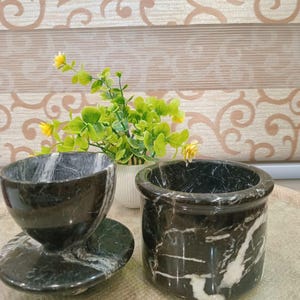 May include: A black marble teacup with saucer and a small cylindrical container. The marble has white veining throughout. A small white vase with greenery and yellow flowers is in the background. The items are on a light brown surface.