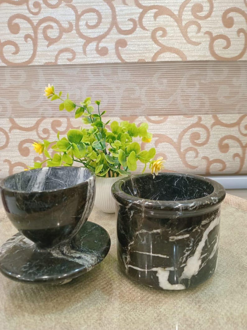 May include: A black marble teacup with saucer and a small cylindrical container. The marble has white veining throughout. A small white vase with greenery and yellow flowers is in the background. The items are on a light brown surface.