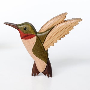 May include: Two wooden hummingbird sculptures. The birds are crafted with natural wood tones, green, red, and brown accents. The wings are spread, showcasing the detailed wood grain. The sculptures are set against a white background.
