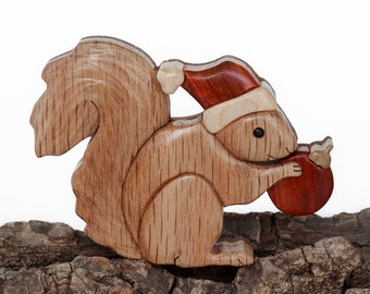 May include: A wooden squirrel figurine with a Santa hat, holding a round nut. The squirrel is made of light and dark brown wood, with a red and white hat. The figurine is placed on a piece of textured bark.