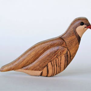 Puede incluir: Una escultura de pájaro de madera tallada en forma de perdiz. El pájaro es marrón con marcas marrón claro y blancas. El pico del pájaro es rojo.