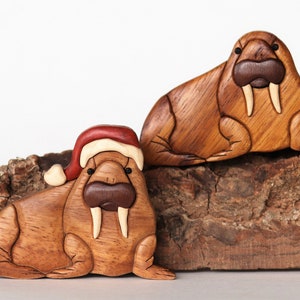May include: Two wooden walrus figurines. One walrus wears a Santa hat. Both have tusks and are made of light and dark brown wood. They are displayed on a piece of textured, brown bark.