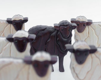 May include: A group of wooden sheep figurines, one dark brown and the others white, against a white backdrop. The sheep have dark brown faces and ears. The white sheep have a light wood grain pattern.