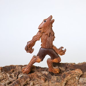 Può includere: Una statuetta di legno di un lupo mannaro, scolpita in legno marrone con accenti marrone scuro. Il lupo mannaro è in piedi sulle zampe posteriori, con la testa inclinata all'indietro e la bocca aperta in un ululato. La statuetta è dettagliata con pelliccia, artigli e denti.