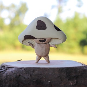Può includere: Una figura di fungo in legno con un cappello color crema con macchie marroni scure. La figura ha una faccia sorridente e braccia tese, in piedi su una base di legno. Un oggetto decorativo affascinante.