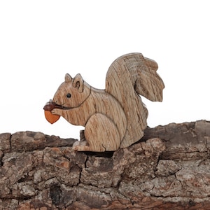May include: A wooden squirrel figurine, holding an acorn, sits on a piece of textured bark. The squirrel is carved from light brown wood, with visible grain details. The acorn is a contrasting shade of brown.