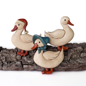 Pode incluir: Tr&ecirc;s figuras de pato de madeira, cada uma com um chap&eacute;u diferente. O pato &agrave; esquerda est&aacute; usando um chap&eacute;u de Papai Noel vermelho, o pato do meio est&aacute; usando um chap&eacute;u azul e o pato &agrave; direita est&aacute; sem chap&eacute;u. Os patos est&atilde;o em p&eacute; em um peda&ccedil;o de casca de &aacute;rvore.