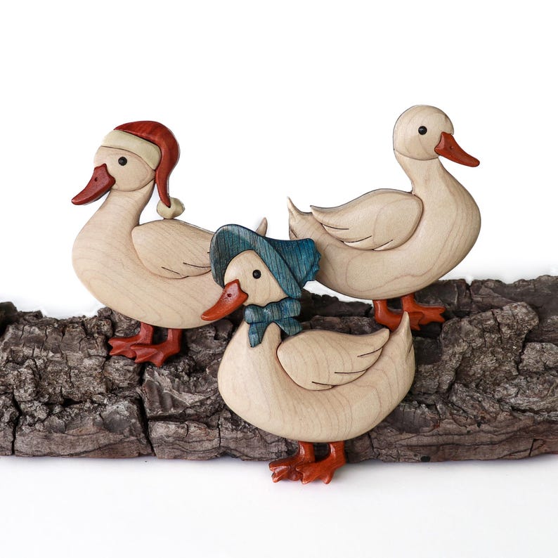 Pode incluir: Tr&ecirc;s figuras de pato de madeira, cada uma com um chap&eacute;u diferente. O pato &agrave; esquerda est&aacute; usando um chap&eacute;u de Papai Noel vermelho, o pato do meio est&aacute; usando um chap&eacute;u azul e o pato &agrave; direita est&aacute; sem chap&eacute;u. Os patos est&atilde;o em p&eacute; em um peda&ccedil;o de casca de &aacute;rvore.