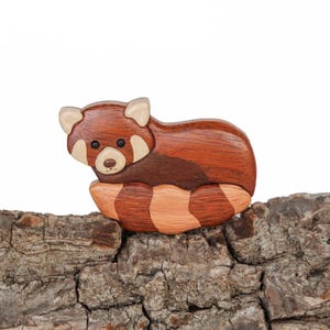 May include: A wooden red panda figurine with a brown and orange striped tail, sitting on a piece of tree bark.