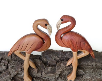 Puede incluir: Dos figuritas de flamenco de madera talladas en madera con una mancha marrón rojiza. Las figuritas se enfrentan y están de pie sobre un trozo de corteza de árbol.