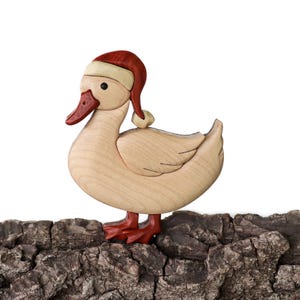 Pode incluir: Um ornamento de pato de madeira usando um chap&eacute;u de Papai Noel vermelho e branco. O pato est&aacute; em p&eacute; em um peda&ccedil;o de casca de &aacute;rvore.