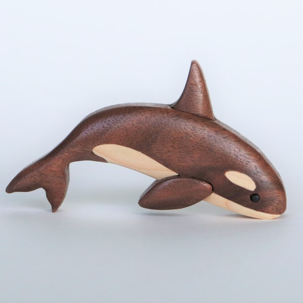 May include: A wooden carved orca whale figurine with a brown body and a white patch on its back. The figurine is painted with black eyes and a white stripe on its side.