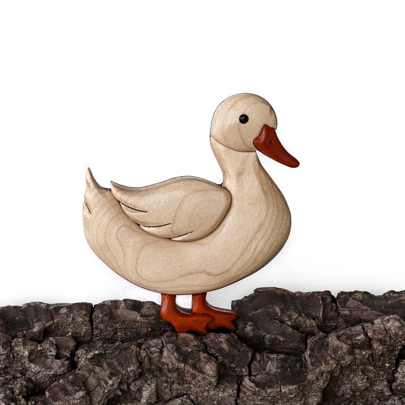 Pode incluir: Uma escultura de madeira de um pato com um corpo castanho claro, um bico e patas laranja. O pato est&aacute; em p&eacute; sobre um peda&ccedil;o de casca de &aacute;rvore.