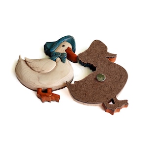 Pode incluir: Uma figura de pato de madeira com um chap&eacute;u azul e um suporte de feltro marrom. O pato &eacute; pintado de branco com detalhes marrons e usa um chap&eacute;u azul e branco. O suporte de feltro &eacute; marrom e tem um &iacute;m&atilde; na parte de tr&aacute;s.