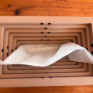 May include: A rectangular, light peach-coloured tissue box cover featuring a nested, tiered frame design. The structure consists of multiple concentric rectangular layers that create a stepped appearance, with a single white tissue emerging from the centre.
