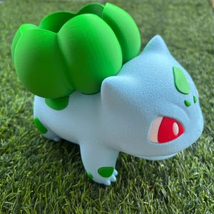 May include: A light blue creature-shaped planter with a textured surface, featuring a large, rounded green bulb-like structure on its back. The figure has pointed ears, red eyes with white highlights, and dark green spots on its body.
