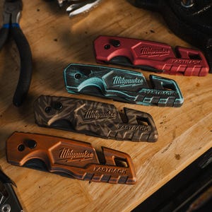 May include: Four Milwaukee Fastback utility knives in red, teal, brown, and orange, with the brand name and "Fastback" text visible. The knives are on a wooden surface, with tools in the background.