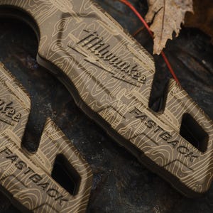 K&ouml;nnte beinhalten: Nahaufnahme eines beigefarbenen Universalmessers mit einem topografischen Kartenmuster. Das Messer tr&auml;gt die Logos "Milwaukee" und "FASTBACK". Der Griff hat ein strukturiertes Design, und ein schwarzer Knopf ist sichtbar. Das Messer liegt auf einer dunklen, strukturierten Oberfl&auml;che.