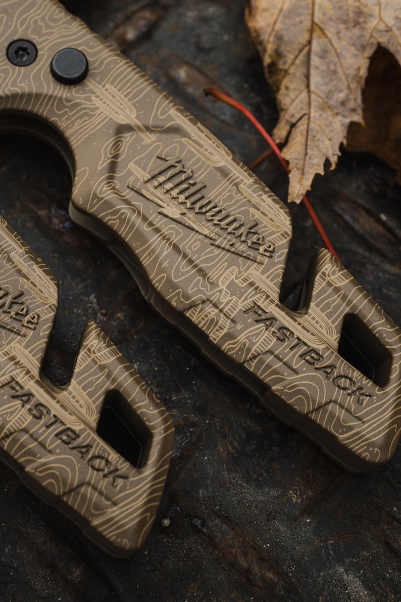K&ouml;nnte beinhalten: Nahaufnahme eines beigefarbenen Universalmessers mit einem topografischen Kartenmuster. Das Messer tr&auml;gt die Logos "Milwaukee" und "FASTBACK". Der Griff hat ein strukturiertes Design, und ein schwarzer Knopf ist sichtbar. Das Messer liegt auf einer dunklen, strukturierten Oberfl&auml;che.