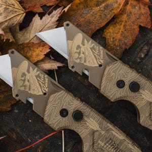 K&ouml;nnte beinhalten: Zwei Gebrauchsmesser mit beigefarbenen Griffen und silbernen Klingen. Die Griffe weisen ein topografisches Kartendesign und eine Totenkopf-Grafik mit dem Text "Hell Raiser" auf. Die Messer liegen vor einem Hintergrund aus Herbstbl&auml;ttern.