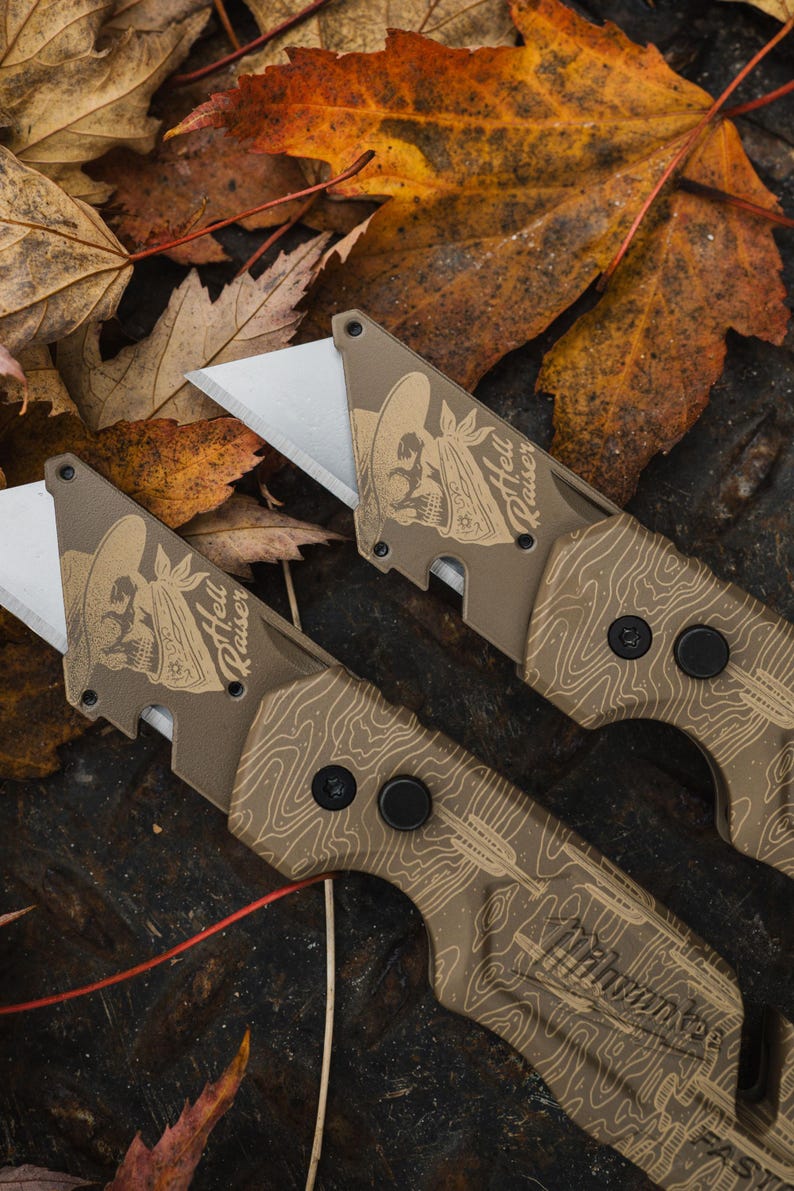 K&ouml;nnte beinhalten: Zwei Gebrauchsmesser mit beigefarbenen Griffen und silbernen Klingen. Die Griffe weisen ein topografisches Kartendesign und eine Totenkopf-Grafik mit dem Text "Hell Raiser" auf. Die Messer liegen vor einem Hintergrund aus Herbstbl&auml;ttern.