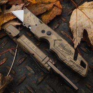 K&ouml;nnte beinhalten: Ein beigefarbenes Universalmesser und seine Scheide werden auf einer Metalloberfl&auml;che mit Herbstlaub pr&auml;sentiert. Das Messer hat eine silberne Klinge und den Text "Milwaukee FASTBACK". Auf der Scheide steht "REBEL TECH".
