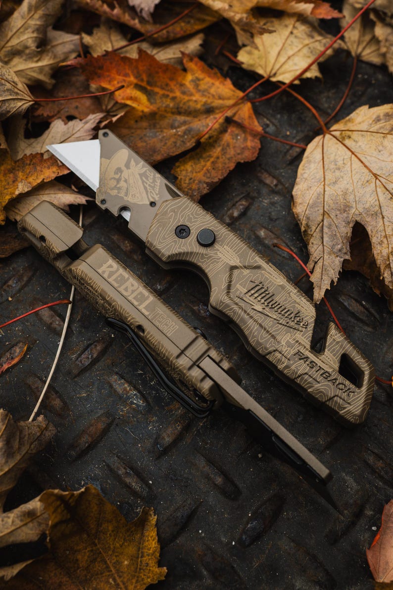 K&ouml;nnte beinhalten: Ein beigefarbenes Universalmesser und seine Scheide werden auf einer Metalloberfl&auml;che mit Herbstlaub pr&auml;sentiert. Das Messer hat eine silberne Klinge und den Text "Milwaukee FASTBACK". Auf der Scheide steht "REBEL TECH".