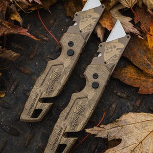 Puede incluir: Se muestran dos cuchillos utilitarios marrones con hojas blancas. Los cuchillos tienen un patrón topográfico y el texto "Milwaukee" y "FASTBACK". Cada cuchillo presenta un gráfico de calavera con el texto "Hell Raiser". Los cuchillos están sobre una superficie metálica con hojas caídas.