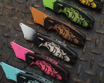 Könnte beinhalten: Eine Auswahl an Gebrauchsmessern mit schwarzen Griffen und Klingen in verschiedenen Farben, darunter Gelb, Grün, Orange, Silber, Rosa und Blau. Jedes Messer hat ein dekoratives Design und den Text "REBEL VISION".