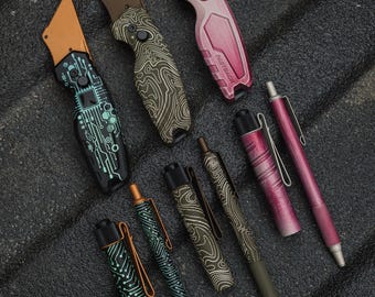Könnte beinhalten: Eine Auswahl an Gebrauchsmessern und Stiften in verschiedenen Farben und Designs. Die Messer haben Metallklingen und gemusterte Griffe. Die Stifte weisen ähnliche Muster und Farben auf, darunter Leiterplatten- und topografische Designs. Die Artikel sind auf einer strukturierten, dunklen Oberfläche angeordnet.