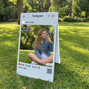 Op de afbeelding: Een witte fotolijst in Instagram-stijl met een persoon erin, zittend op het gras in een park. De lijst bevat het Instagram-logo, like-, commentaar- en deel-pictogrammen en een QR-code. De tekst op de lijst luidt "foreis_yoga" en "yoreis_yoga".