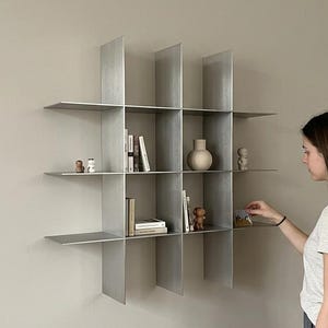 May include: A wall-mounted metal shelving unit featuring a grid design with four vertical dividers and three horizontal shelves, creating twelve open compartments. The shelves are finished in a brushed silver tone and hold various small decorative items.